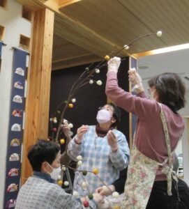 エプロンをつけた3人の女性が、長い枝に色とりどりの団子飾りを取り付けている様子を捉えた写真