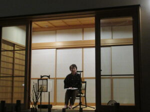 和室の縁側に置かれた椅子に作務衣姿の女性が座り、開いたガラス戸の外に向かって、マイクを使って話をしている様子を正面から撮影した写真