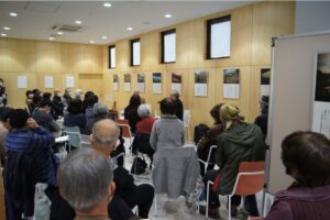 広い室内で多くの来場者が前方の展示作品や話者に注目しながら椅子に座って静かに鑑賞している様子を捉えた写真