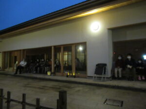 建物の犬走に並んだ椅子に参加者たちが座り、外壁の丸い照明が暗い屋外で明るく光を放っている様子の写真