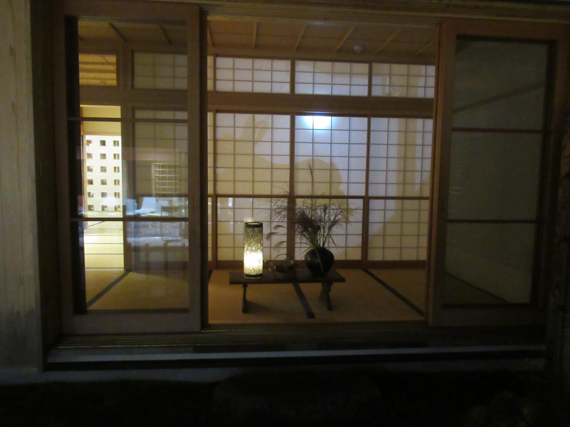 月の明かりとウサギのシルエットが浮かんだ障子の前の畳の上に置かれた台に、行灯とススキなどが飾られた花瓶が置かれている様子を屋外から撮影した写真