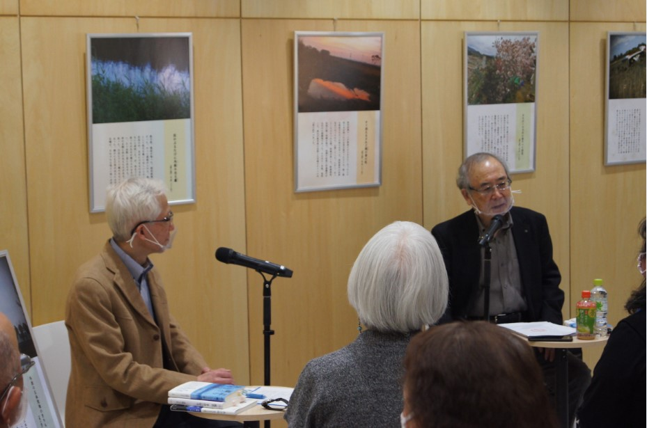 木製パネルに写真作品が展示された室内で二人が卓上のマイクを前に向かい合い、聴衆が静かに見守る様子を捉えた写真