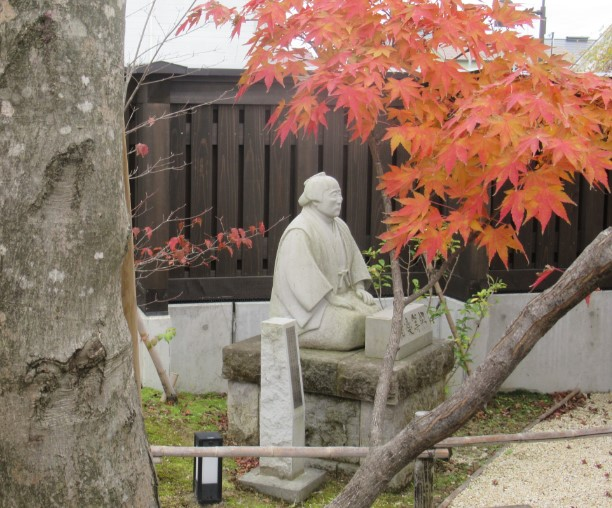 木製の囲いの前で、石の台の上に正座する相楽等躬の石像を、赤く色づいた紅葉の枝越しに撮影した写真
