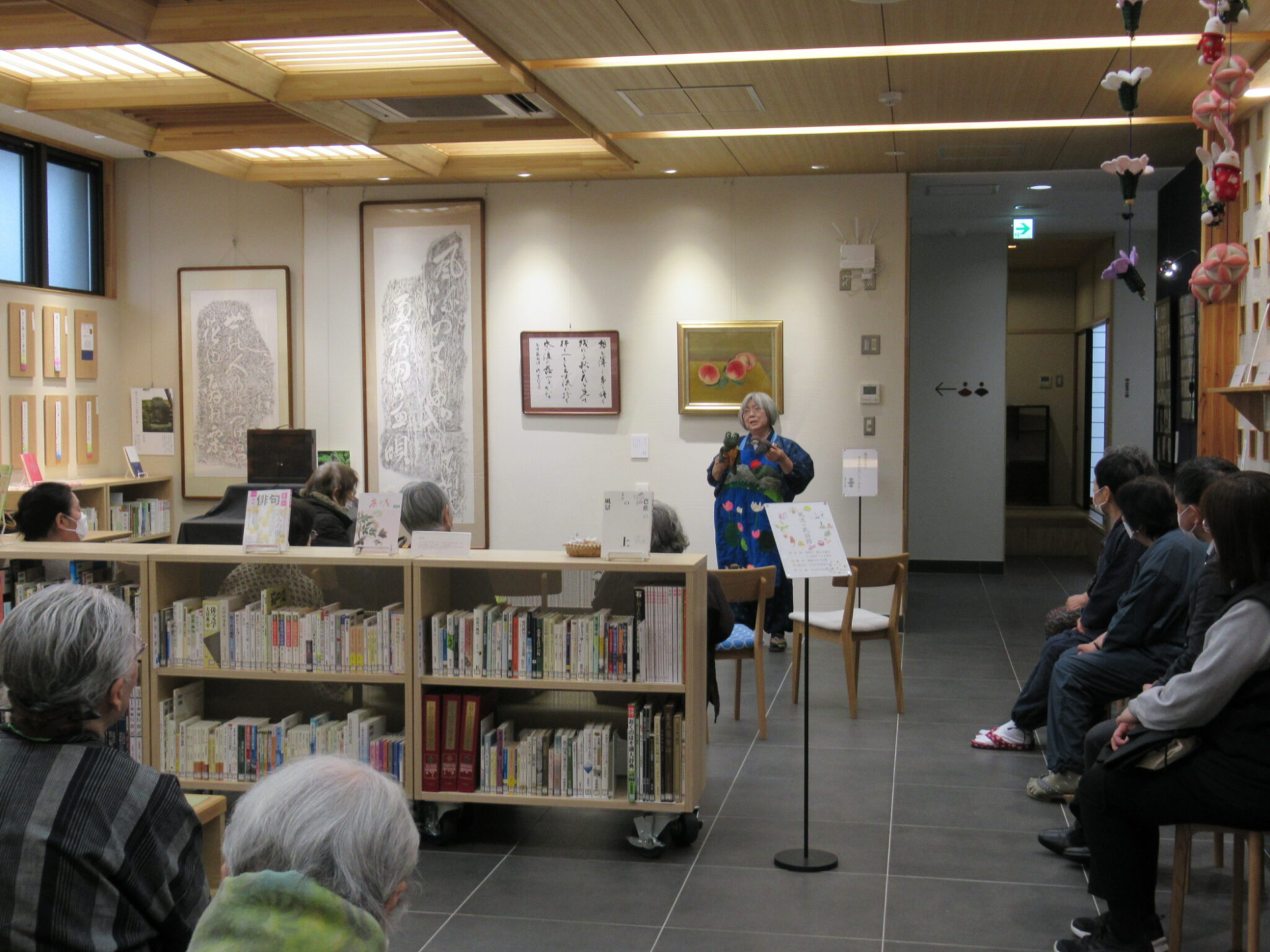 低い本棚が置かれたスペースに並んだ椅子に参加者が座り、絵画や書が飾られた壁の前に立った和装の語り部の女性の話を熱心に聞いている様子の写真