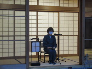和室の障子を背に、作務衣姿の女性が椅子に座り、マイクを使って話をしている写真