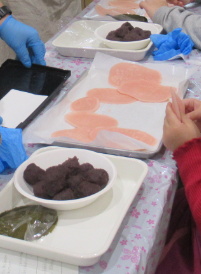 テーブルの上の白い皿に丸めたあんこや桜の葉、白いシートの上に焼きあがった薄い桃色の生地が並んだ写真