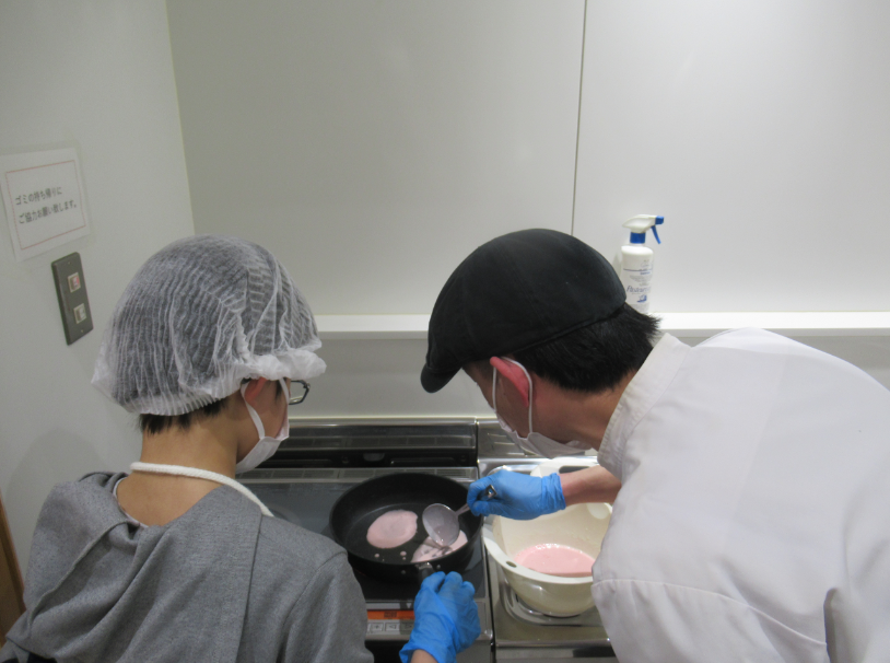 ヘアネットをかぶった参加者と帽子をかぶった講師の男性がフライパンで薄い桃色の生地を焼いている様子の写真