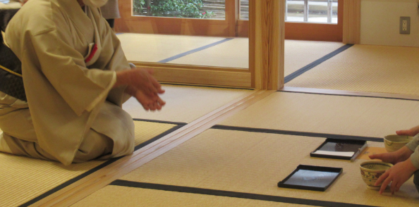 畳の上で着物姿の女性と対面して座った2人の子どもが、抹茶が入った茶碗に手を伸ばしている様子の写真