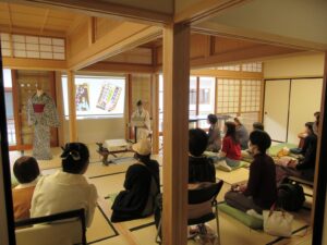和室の奥の壁に資料が映し出され、左に着物を着つけしたマネキン、右に講師の女性が座り、椅子や座布団に座った参加者たちが話を聞いている様子を部屋の後方から撮影した写真