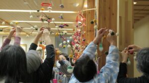 複数の女性たちが高い枝に色とりどりの団子飾りを協力して取り付けている様子を写した写真