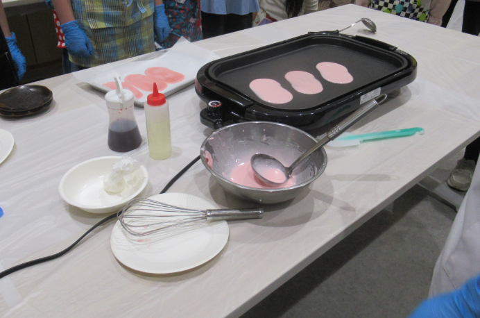 ボウルや泡立て器などの道具が並んだテーブルの上に置いたホットプレートで、ピンク色の生地が楕円形に焼かれている様子を写した写真