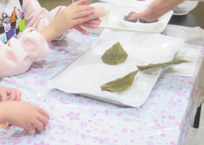 白いシートの上に並んだ桜の葉を前に、子どもたちがピンク色の生地であんこを包んでいる様子の写真