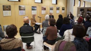 写真展の作品が並ぶ室内で多くの聴衆が椅子に座り前方の対談者を静かに見守る様子を捉えた写真