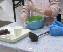 まるめたあんこが入った皿やお玉が置かれた作業台の上で、ボウルに入った生地を泡だて器で混ぜる手元の写真