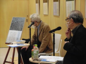 室内で写真パネルが並ぶ壁の前に二人が資料や飲み物を置いた机を挟んでマイク越しに向き合って座る光景の写真