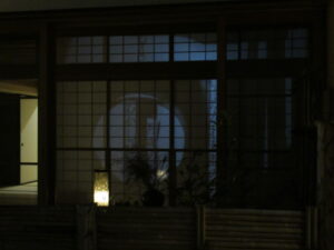 夜の静かな屋外から障子越しに室内を望むと大きな円形の灯りが柔らかく投影され、建物の外壁や手前の植物の影が淡く浮かび上がっている写真