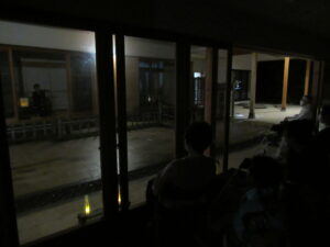 暗い室内に座った参加者たちが、ガラス戸越しに雨の中庭を挟んで、向かいの建物の縁側に座る話者の話を聞いている様子の写真