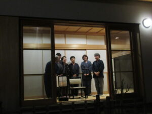 夜の外側から明るい和室を望み、作務衣姿の複数の人物が笑顔で横一列に並んでいる様子を写した写真