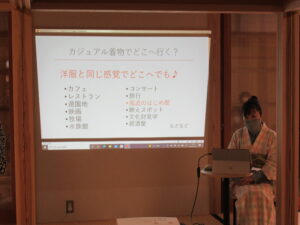 着物姿の女性が資料を写した壁の横に座って話をしている様子の写真