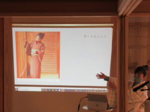 着物姿の女性の資料が映し出された壁を指し示しながら、講師の女性が説明をしている様子を捉えた写真