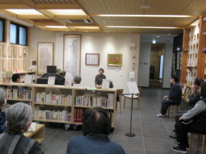 低い本棚を囲むように配置された椅子に参加者が座り、絵画や書が飾られた壁の前に立つ語り部の女性に注目している様子の写真
