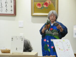 絵画や書が飾られた壁の前に立った、池や山が表現されたエプロンをつけた語り部の女性が、カエルとスズメの人形を両手に持ち話をしている写真