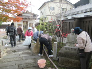 葉が赤く色づいた木がある庭園で、ほうきやごみ袋を持った複数の男女が石畳の通路や植栽の清掃を行い、手前では女性が相楽等躬の石像の清掃を行っている様子の写真