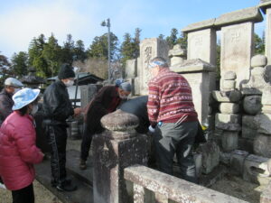 古い石碑が並ぶ前で、数人の男女が集まってお参りをしている様子の写真