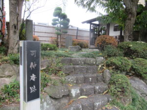 「可伸庵跡」の碑の右側に、低木の植栽に囲まれた数段の石段があり、石段の上には白い壁の建物が佇んでいる写真