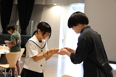 白いセーラー服姿の女子学生が笑顔を浮かべながら、黒い服を着た人物と向き合い、手元で何かを丁寧に受け渡し、交流を行っている写真