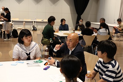 屋内のイベント会場で、白いセーラー服の女子学生やボーダー柄の少年のグループの席に、スーツを着た年配の男性が一緒に座り、身振り手振りを交えながら熱心に語りかけているワークショップの写真