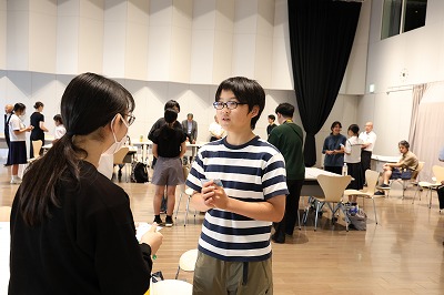 屋内の広いホールのような場所で、ボーダー柄のTシャツを着た眼鏡の少年が、手元のカードを手に持ちながら対面にいる女性へ真剣な表情で話しかけている写真