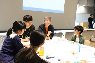 屋内の明るい照明が灯る広い会場で、プロジェクターをバックに、テーブルを囲んだ学生たちのグループの中に大人が一緒に入り、話し合っている写真