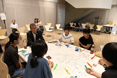屋内の明るいホールで、スーツ姿の年配の男性が笑顔で学生たちと机を囲み、模造紙にカラーペンで図解やアイデアを書き込みながら、和やかな雰囲気の中で活発に意見を交わしているワークショップの様子を捉えた写真