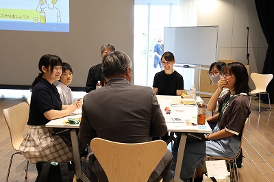 屋内の明るいホールで、スーツ姿の男性を囲んだ学生たちがテーブルにつき、和気あいあいとした雰囲気の中で互いに顔を見合わせて笑顔で意見を出し合っている様子を捉えた写真