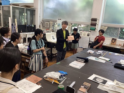 実習室のような室内で、黒いジャケットを着た男性講師が手に持った作品を提示しながら説明し、その話を真剣に聞いている学生たちの写真