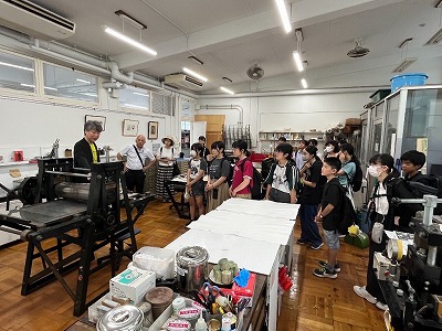 作業室のような場所で、たくさんの学生のグループが講師の説明を真剣な表情で聞いている写真