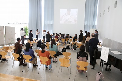 明るい大ホールで多くの参加者が椅子に座り、前方の白い壁に投影されたスクリーンに映像が映し出されている写真