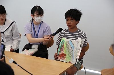 縞模様のシャツを着た少年がカラフルな絵が描かれたスケッチブックを広げて持ち、その口元に眼鏡をかけた女性がマイクを差し出して発表をサポートしている写真