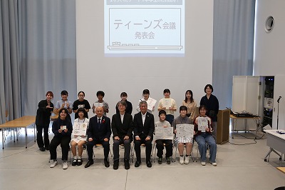 「ティーンズ会議発表会」とスクリーンに映し出された白い壁を背景に、賞状や作品を手にした若者たちとスーツ姿の男性たちが笑顔で並んでいる写真