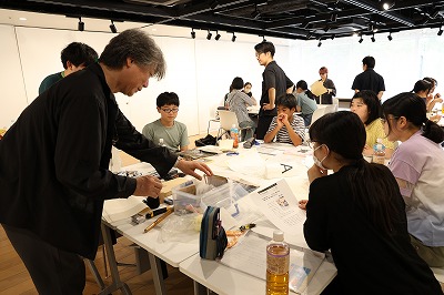 講師の男性がテーブルの上の箱から道具を取り出しながら熱心に説明を行い、それを取り囲むように座っている学生たちが真剣な表情で見ている写真