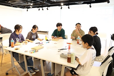 明るい照明が灯る室内の白いテーブルを囲み、若者たちと大人の男性がリラックスした表情で座り、一人の少年が手を挙げて発言する様子を捉えた写真