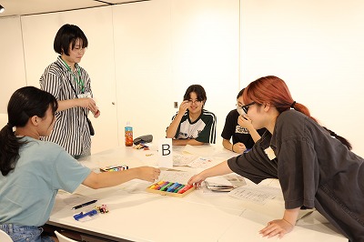 屋内の白いテーブルを囲み、ネームプレートを下げた私服姿の若者たちが、カラーペンを手に取りながら模造紙に意見を書き込み、身を乗り出して互いのアイデアを熱心に共有し合っている写真