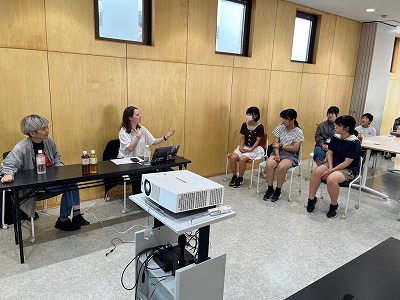 木目調の壁が印象的な明るい室内で、ノートパソコンを開いた女性講師が身振り手振りを交えて熱心に話し、その前で椅子に座った学生たちが真剣な表情で耳を傾けている写真