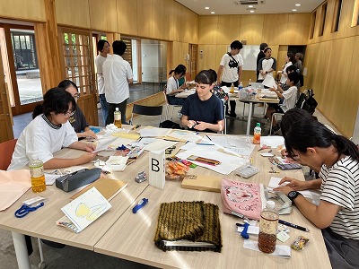 木目調の壁が広がる明るい室内で、複数の男女が机を囲んで紙を広げたり作業をしたりしながら交流しているワークショップを捉えた写真