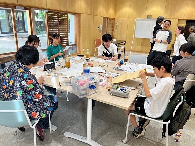 木目調の壁に囲まれた明るい室内で、子供から大人までが大きなテーブルを囲み、紙や道具を使って真剣に作業をしたり談笑している様子を捉えた写真