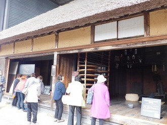 歴史を感じさせる茅葺き屋根の古民家の前に、参加者たちが土間や囲炉裏などの内部の様子を熱心に覗き込んでいる写真