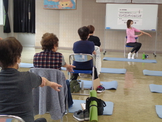 室内で、ホワイトボードの前に座るピンク色のウェアを着た講師の女性の動きを参考にしながら、手前にヨガマットを敷き、椅子に座った参加者たちが片腕を伸ばして筋肉をほぐすリンパコンディショニングを行っている様子の写真