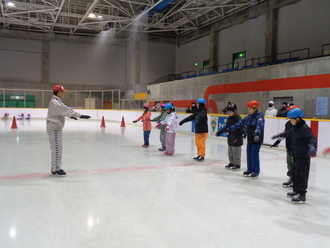 広々としたアイススケートリンクの上で、色とりどりのヘルメットを被った子どもたちが一列に並び、前方に立つ指導者の動きに合わせて腕を伸ばしながら基本姿勢を教わっている様子の写真