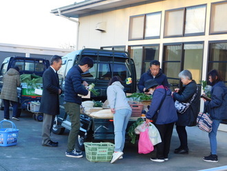 屋外で、2台の軽トラックの荷台に積まれた新鮮な大根や葉物野菜を囲み、地域の人々が品定めをしたり袋に詰めたりしながら買い物を楽しんでいる様子を写した写真